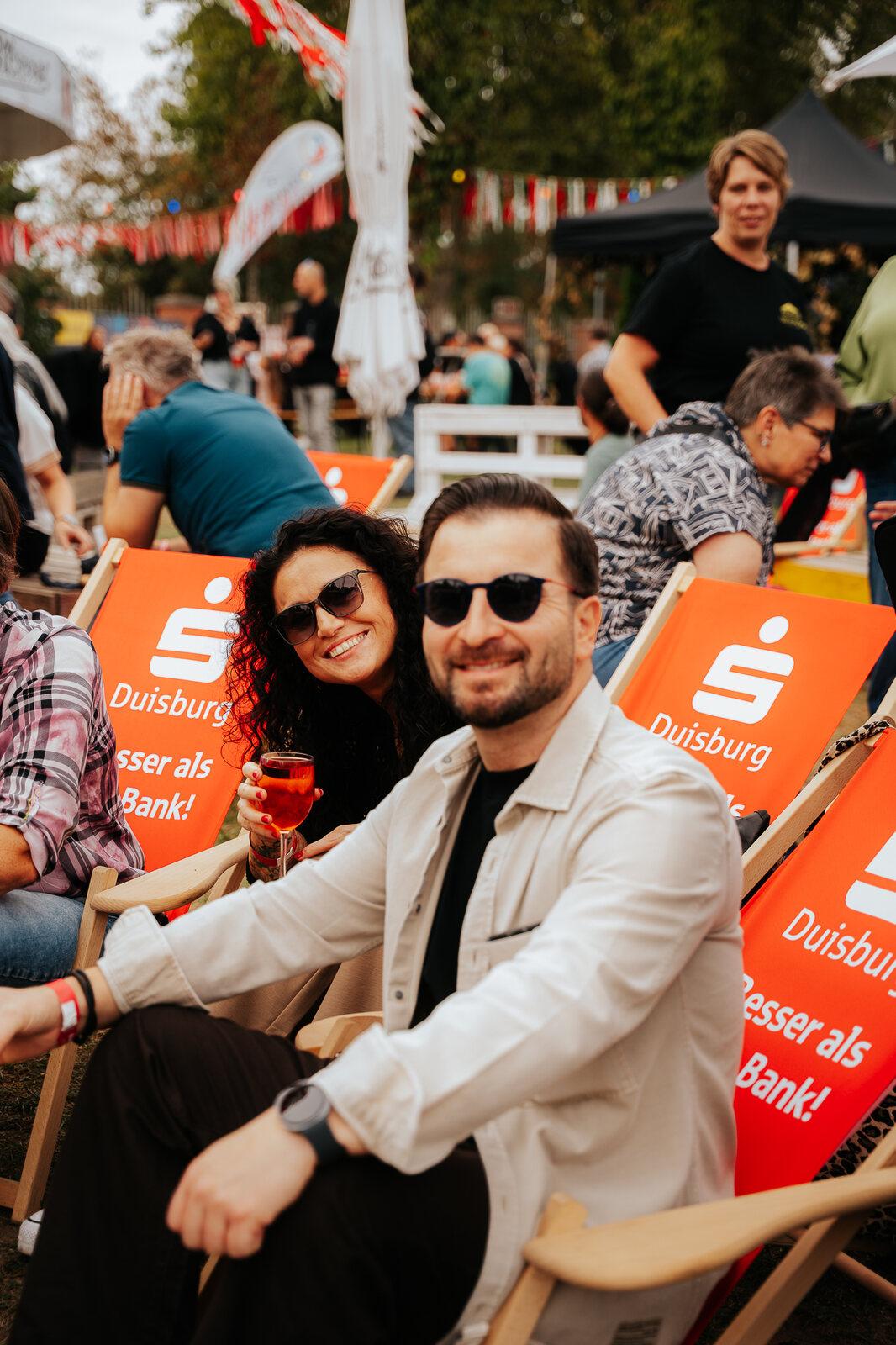 Ein Paar mit Sonnenbrillen sitzt entspannt in orangefarbenen Liegestühlen und stößt mit einem Getränk an