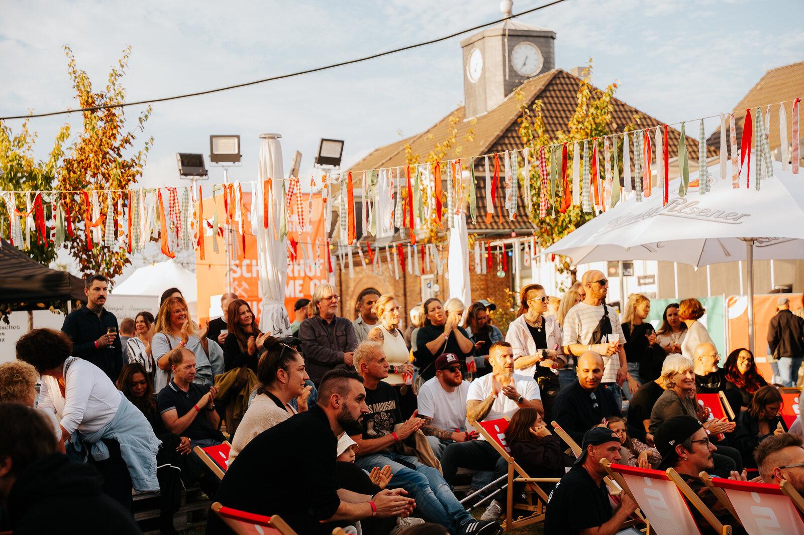 Applaudierendes Publikum im Lounge- und Biergartenbereich unter bunten Wimpelketten und Sonnenschirmen