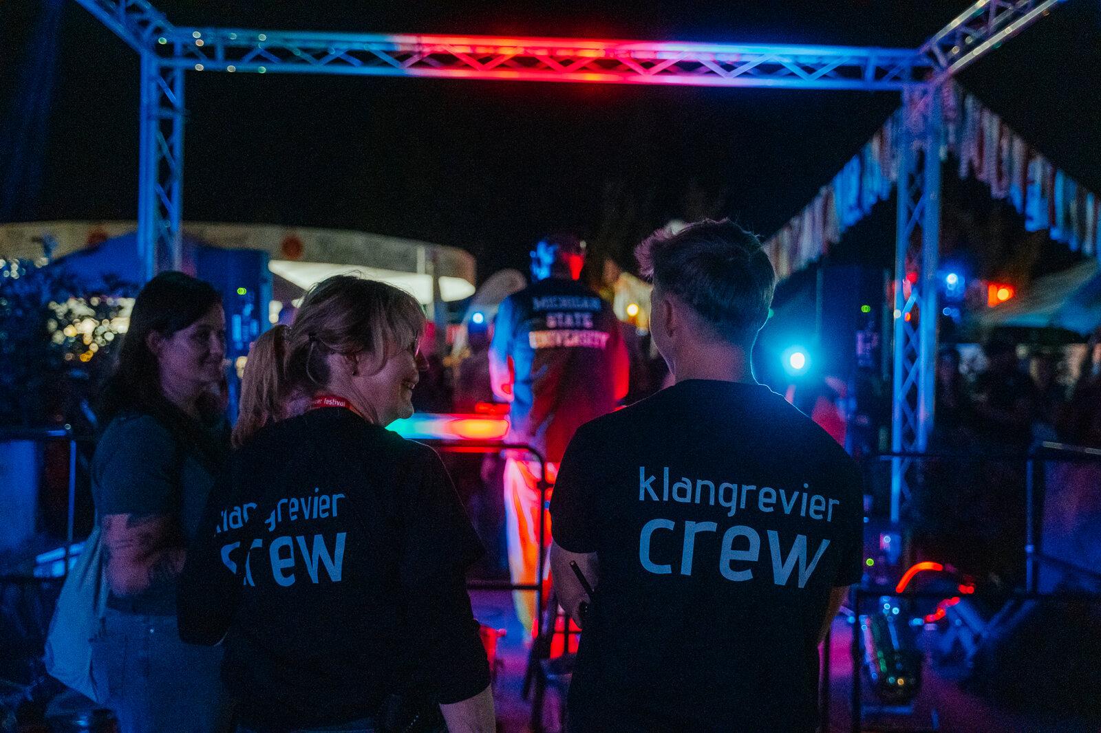 Zwei Helferinnen und Helfer in klangrevier-crew-Shirts am Abend vor der blau und rot ausgeleuchteten Bühnentraverse
