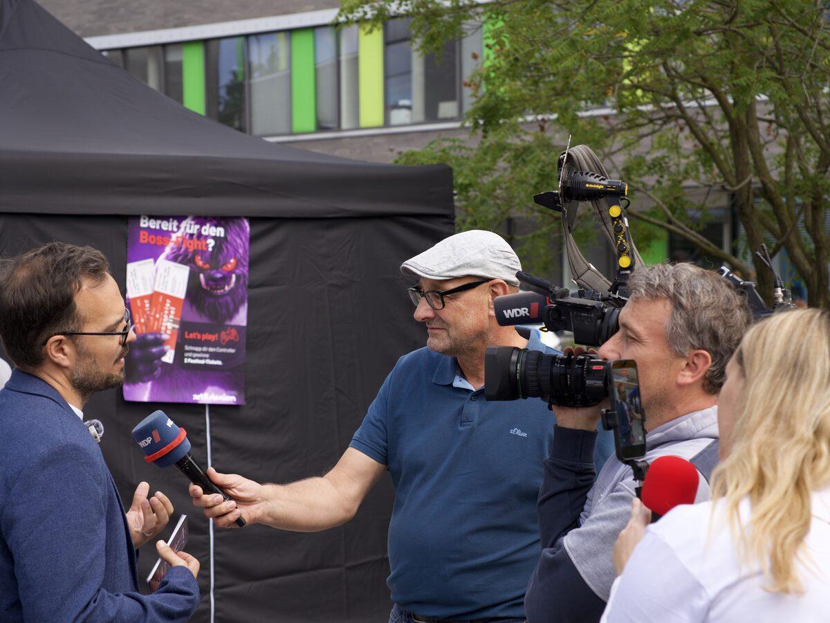 Ein WDR-Team filmt am Messestand von art&design ein Interview, im Hintergrund das Plakat zum Zukunftsschreck-Spiel