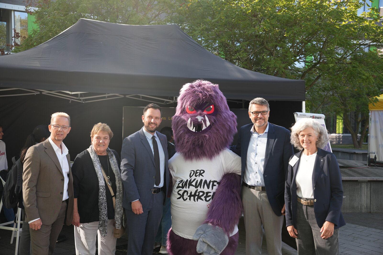 Vertreterinnen und Vertreter der beteiligten Städte gemeinsam mit dem Maskottchen DER ZUKUNFTSSCHRECK vor dem Messezelt auf dem Campus
