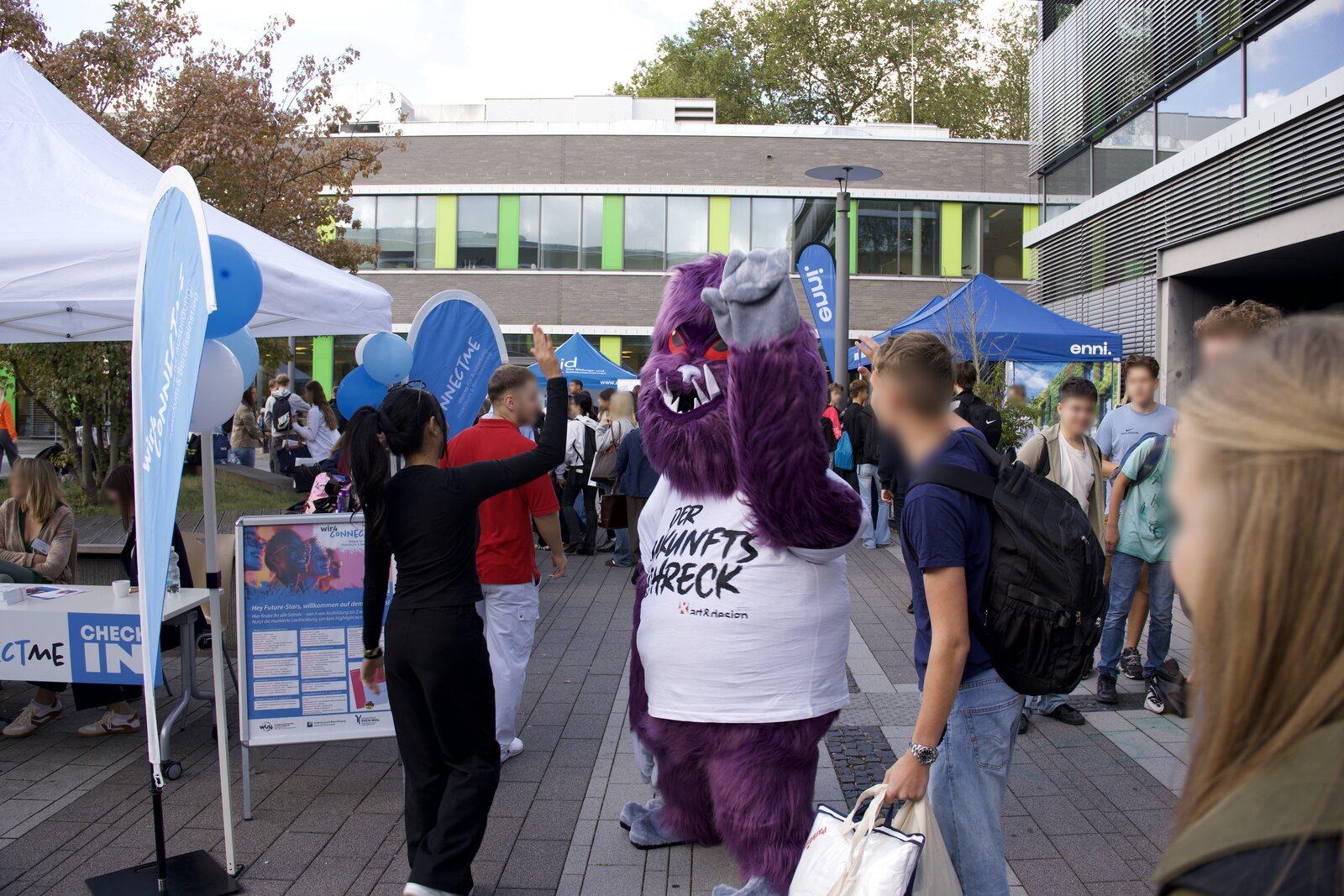 Das Maskottchen DER ZUKUNFTSSCHRECK zwischen Besucherinnen und Besuchern vor den Zelten am Check-in der wir4connectMe