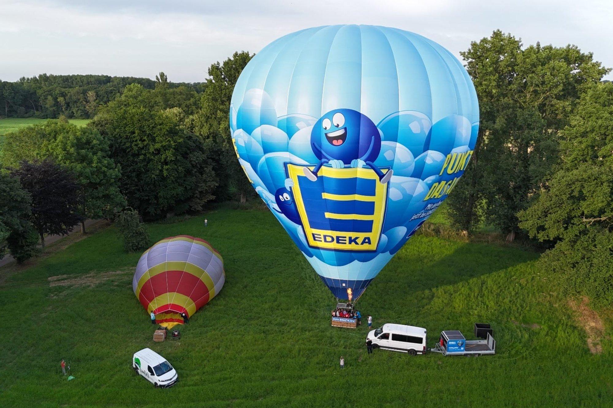 EDEKA-Heißluftballon beim Start auf einer Wiese, daneben ein weiterer Ballon und Begleitfahrzeuge