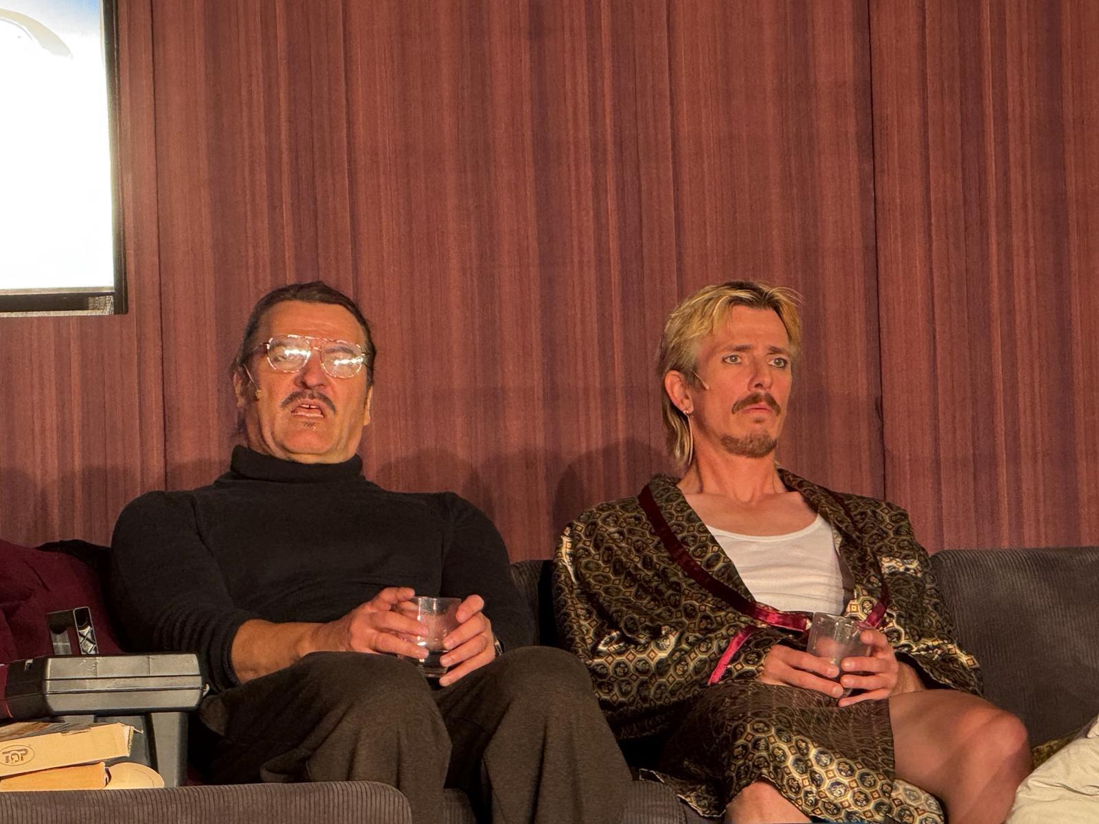 Zwei Darsteller sitzen mit Gläsern in der Hand auf dem Sofa