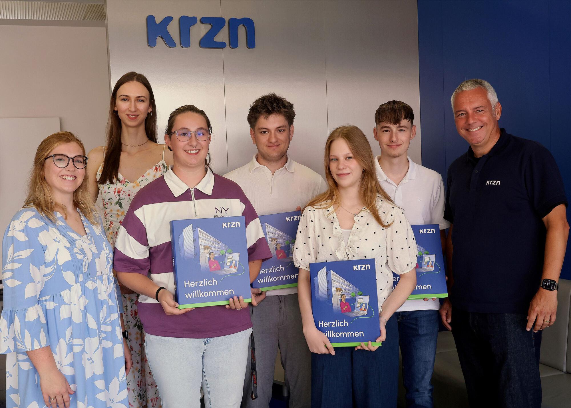 Die drei neuen Auszubildenden und die Studentin des KRZN mit ihren Ausbildungskoordinatorinnen und dem Geschäftsleiter vor der krzn-Wand