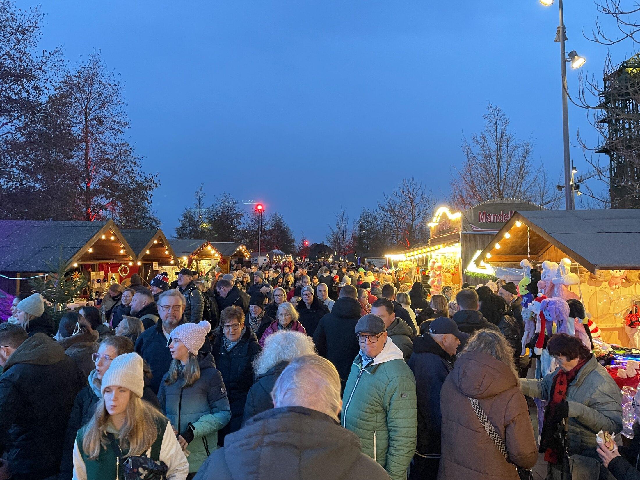 Dicht gefüllte Marktgassen auf dem Weihnachtsmarkt im Zechenpark zur blauen Stunde, dahinter beleuchtete Hütten und der Förderturm