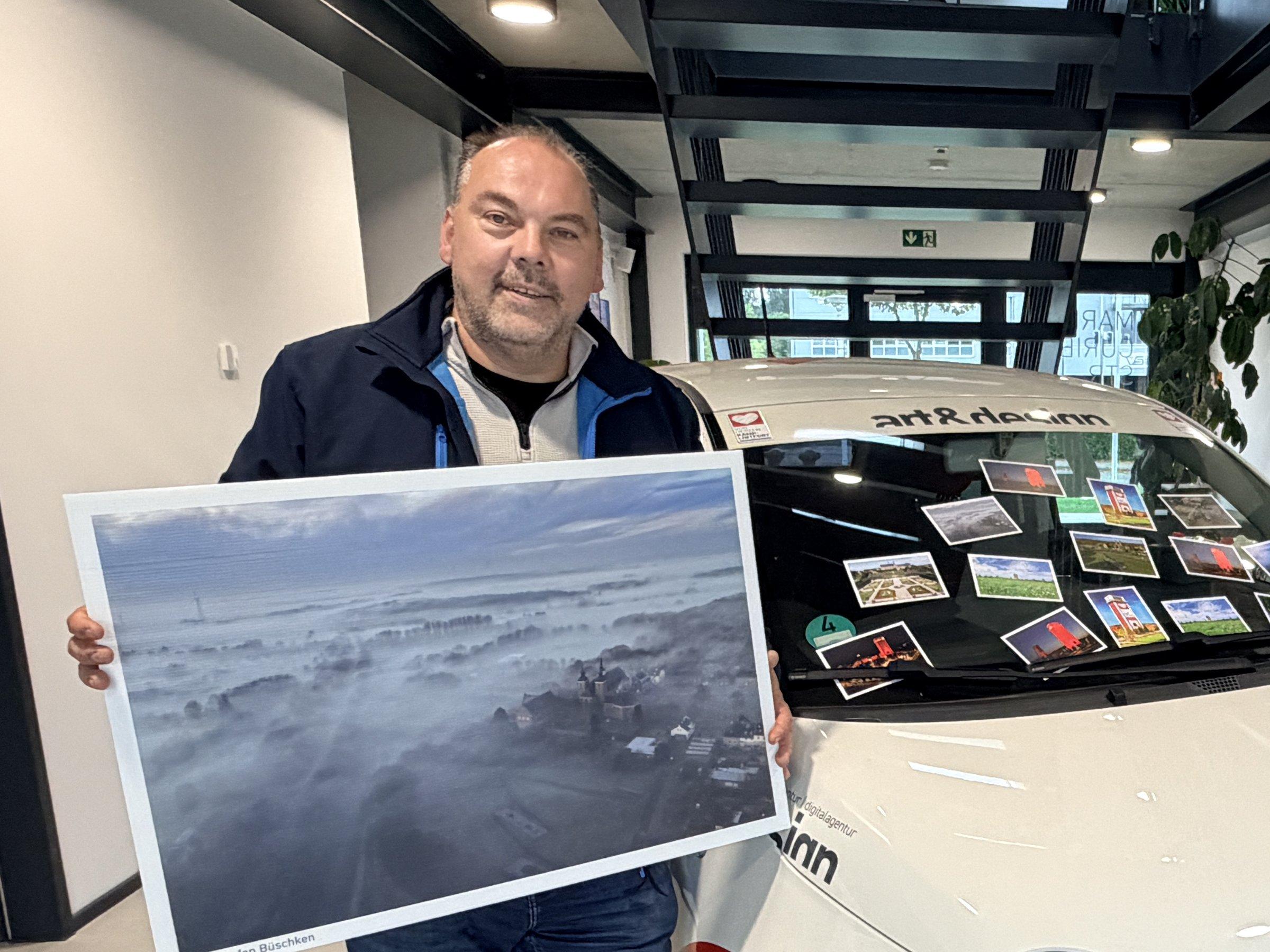 Fotograf Stefan Büschken mit einem großformatigen Nebel-Luftbild von Kamp-Lintfort vor dem art&design-Auto, auf dessen Windschutzscheibe die zehn Postkartenmotive liegen