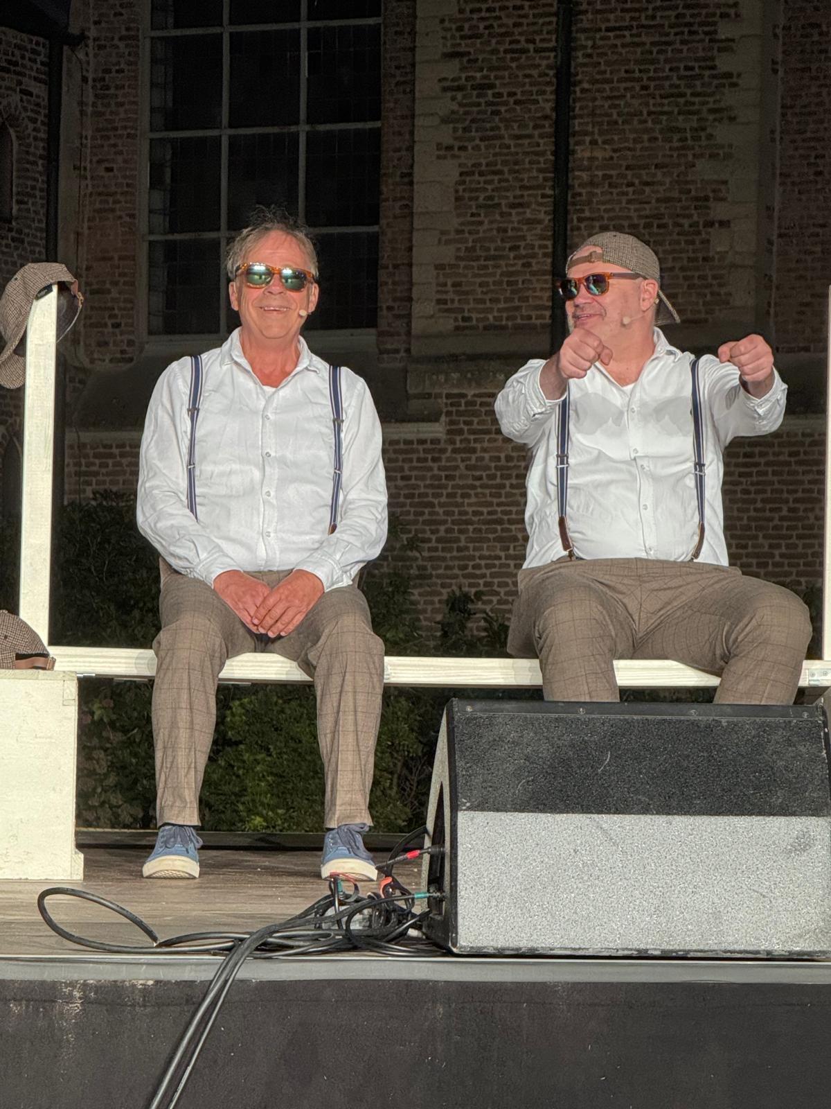 Zwei Darsteller aus „König Ödipus“ in weißen Hemden mit Hosenträgern sitzen auf der Bühne vor der Abteikirche